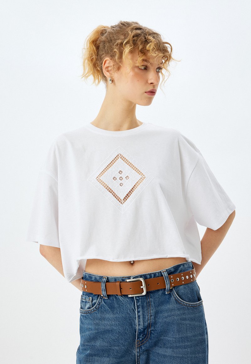 Hvid cropped t-shirt lavet af bomuld, med et diamantformet udskæringsdesign og små udsmykninger. Sammen med blå jeans og et brunt bælte.