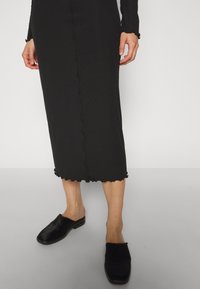 Partie inférieure du corps d'une personne portant une jupe midi noire côtelée avec des bords froncés et des chaussures noires sans lacets sur un fond uni.