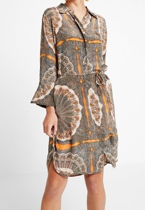 Femme portant une robe-chemise mi-longue avec un motif cachemire en orange, marron et beige, dotée de manches longues et d'une ceinture à la taille.