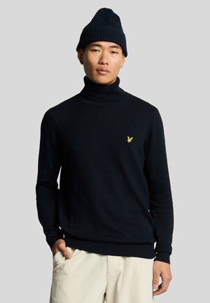Man draagt een zwarte coltrui met een geel logo, een zwarte gebreide muts, crèmekleurige broek en zilveren ringen aan zijn rechterhand.