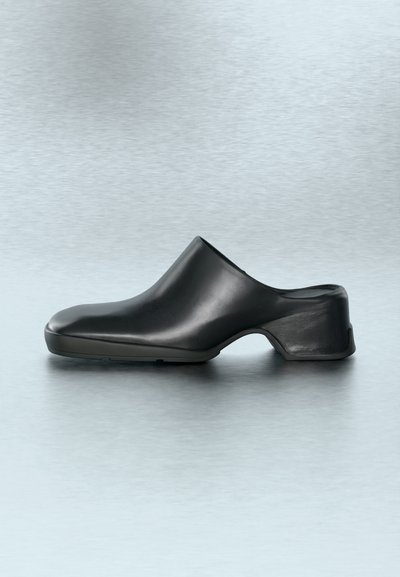 Μαύρο δερμάτινο mule slip-on με χαμηλό τακούνι τύπου block και στρογγυλεμένη μύτη σε λεία γκρι επιφάνεια.
