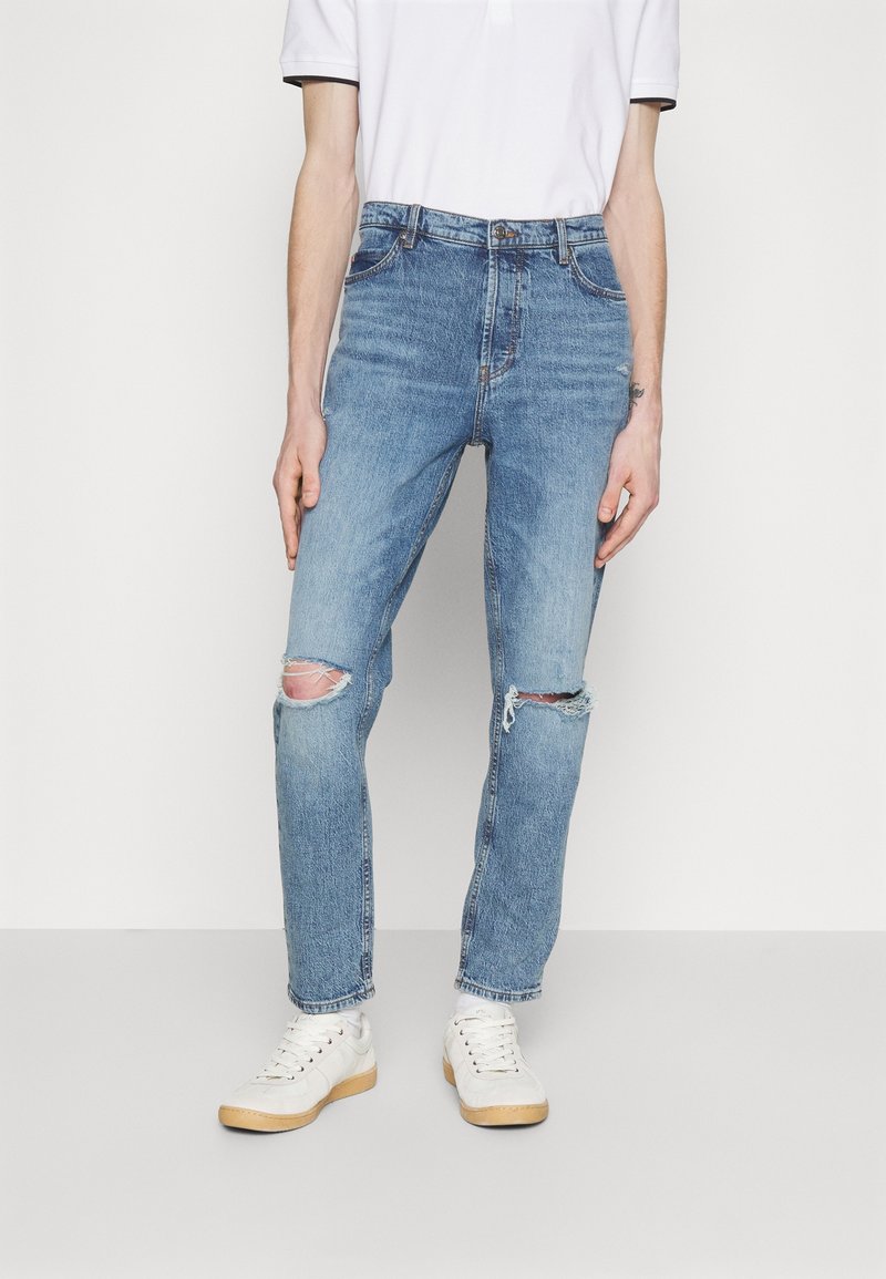 Jean en denim bleu clair déchiré avec bords effilochés, forme fuselée, cinq poches, et fermeture standard à bouton, porté avec des baskets blanches.