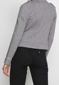 Giacca grigia tagliata con colletto, spalline e finiture dorate sui polsini. Indossata sopra pantaloni neri con etichetta del logo visibile.
