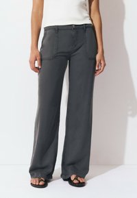 Person wearing dark gray wide-leg pants with front patch pockets and black studded open-toe sandals, standing against a white background.