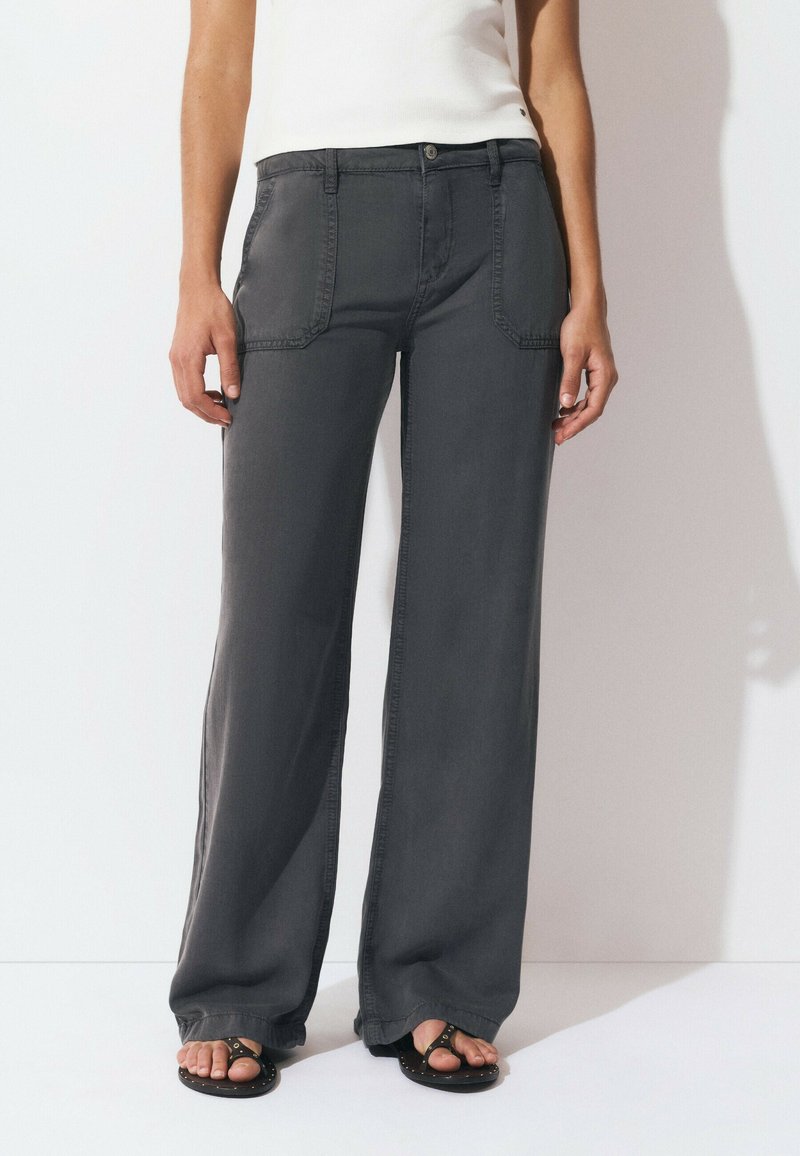 Person wearing dark gray wide-leg pants with front patch pockets and black studded open-toe sandals, standing against a white background.