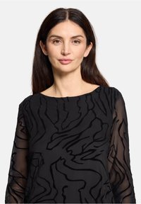 Czarna bluzka z przezroczystymi rękawami, wyszywana teksturowanym falistym wzorem. Dekolt jest okrągły, a tkanina ma gładkie wykończenie.