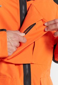 Orange waterproof jacket with a black zipper and accents. Shows a hand opening a pocket with a zip closure, highlighting the interior lining.