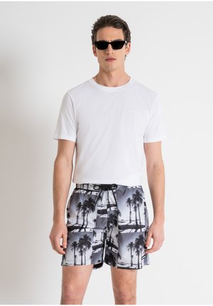 Camiseta blanca de cuello redondo combinada con pantalones cortos de baño de estampado de palmeras en blanco y negro. Los pantalones cortos cuentan con una cintura elástica y cordón ajustable.