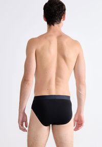 Mann, der mit dem Rücken zur Kamera steht und schwarze Unterhosen trägt, vor einem einfachen weißen Hintergrund.