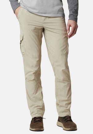 Mann trägt beige Cargo-Hosen und braune Wanderschuhe und steht mit den Händen in der Nähe der Taschen vor einem einfarbigen Hintergrund.