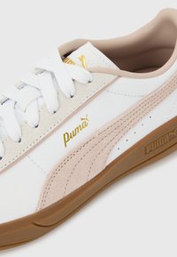 Λευκό και μπεζ αθλητικό παπούτσι Puma με λευκά κορδόνια, χρυσό λογότυπο Puma, σουέτ ρίγα και καφέ λαστιχένια σόλα σε φωτεινό φόντο.