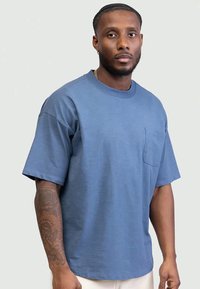 Man met kort haar en baard, gekleed in een blauwe overhemd met korte mouwen en een zak, met een getatoeëerde linkerarm, staand voor een effen achtergrond.
