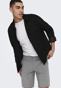 Sort zip-up jakke med en tekstureret overflade og to brystlommer, parret med grå skræddersyede shorts. Modellen er i en dynamisk positur.