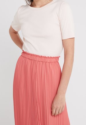 Vrouw draagt een wit shirt met korte mouwen, ingestopt in een hoog taille koraalroze geplooide rok met een gerimpelde tailleband.