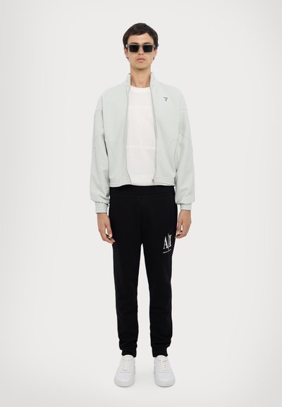 Svetlo šedá bunda na zips, biele tričko, čierne tepláky, biele tenisky; slnečné okuliare dotvárajú look. Logo na teplákoch je viditeľné.