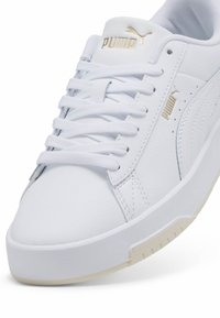 Valge nahkne sneaker tekstureeritud külgedega, lame pael ja kuldne bränding, kerge beež kummist tald. Omab ümarat varbajoont.
