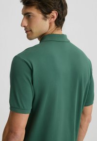Polo shirt verde a maniche corte con un tessuto testurizzato, caratterizzato da un colletto classico e linee pulite, mostrato da dietro.