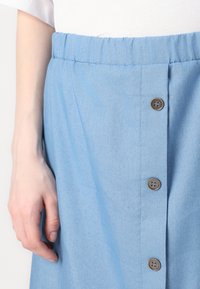 Jupe bleu clair avec une taille élastique froncée, fermeture à boutons à l'avant comportant des boutons marron, et un tissu doux et texturé.