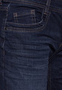 Jean en denim bleu foncé avec une poche arrière dotée de coutures et d'une texture subtile, mise en valeur par un fil orange contrastant.