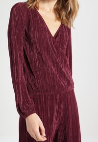 Burgundy pleated jumpsuit with a wrap v-neck and long sleeves, featuring textured fabric and elasticized waist details.