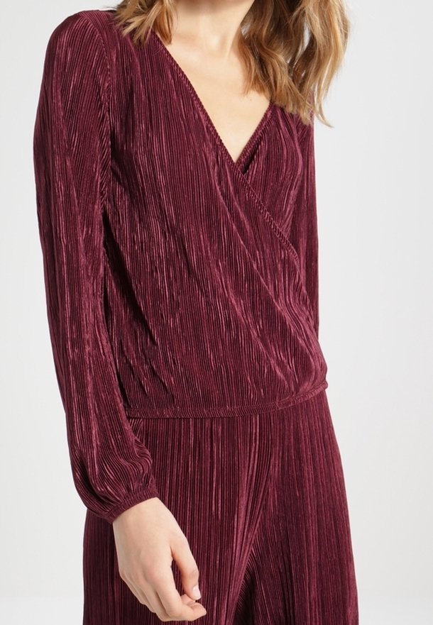 Burgundy pleated jumpsuit with a wrap v-neck and long sleeves, featuring textured fabric and elasticized waist details.