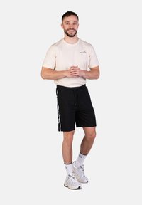 Beige T-Shirt mit Logo, schwarze Sportshorts mit weißen Seitenstreifen, kombiniert mit weißen Sneakers und Sportsocken.