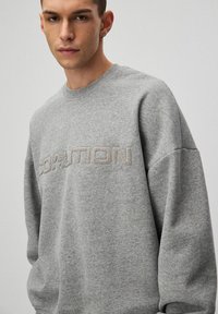 Młody mężczyzna w szarym swetrze z haftem "COALITION" na piersi, stojący na jednolitym jasnym tle.