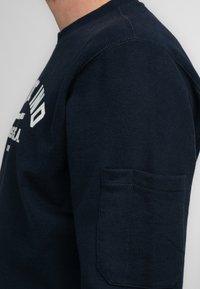 Marineblauwe hoodie met geribbelde kraag, witte logotekst en een zijzakdetail. Gemaakt van zachte katoenen stof met een reguliere pasvorm.