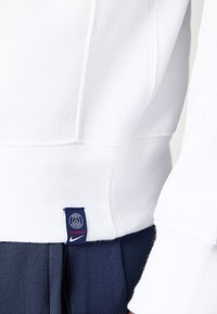 Sweatshirt blanc avec une texture côtelée, arborant une étiquette logo Paris en bleu au bas et un marquage Nike. Tissu navy visible en dessous.