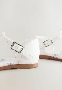 Weiße Sandalen mit einem synthetischen Lederoberteil, verstellbarem Knöchelriemen mit einer Metall-Schnalle und blumenmustergefütterter Innensohle. Niedriges Absatzdesign.