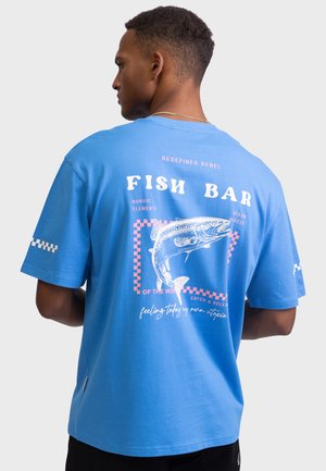 Maglietta blu in cotone con grafica di un pesce, testo in bianco e rosa, e dettagli a scacchi sulla manica sinistra. Vestibilità rilassata, maniche corte.