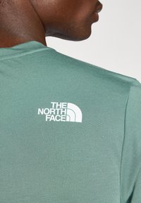 Hele roheline ümmarguse kaelusega T-särk ribatekstuuriga ja valge logo ülaservas, millel on kõver kujunduselement.