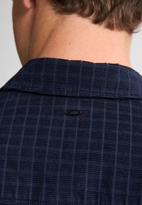 Heren overhemd in marineblauw, met een gestructureerde stof en een subtiel ruitpatroon; beschikt over een klassieke kraag en een klein zwart logotag aan de achterkant.