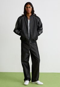 Chaqueta bomber negra con rayas grises, combinada con jeans negros holgados y zapatillas blancas. El modelo está de pie contra un fondo sencillo.