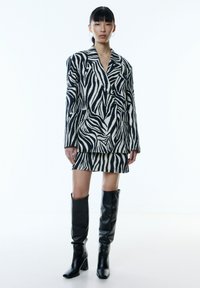 Blazer en mini-rok met zebra-prints in zwart en crème, gecombineerd met hoge zwarte leren laarzen. Gestructureerd ontwerp met lange mouwen.