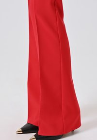 Gambe inferiori che indossano pantaloni larghi rossi che coprono scarpe nere a punta con punte in metallo dorato su uno sfondo chiaro.