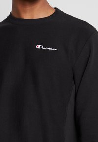 Felpa nera a girocollo con piccolo logo Champion ricamato in bianco e rosso sul lato sinistro del petto, indossata da una persona mostrata dalla scollatura fino a metà torso.