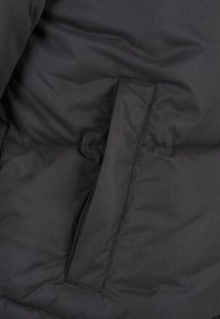 Tissu de veste matelassée noire avec une poche verticale zippée partiellement couverte par un rabat en tissu.