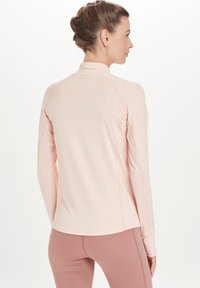 Helles pinkes Langarm-Sportoberteil mit Raglanärmeln, weichem Textil und reflektierenden Akzenten entlang der Schulternaht und des Halses.