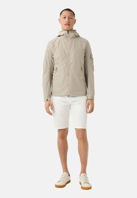 Lightweight, beige jacket with a hood, featuring zippered pockets and a smooth texture. Paired with white shorts and light sneakers.