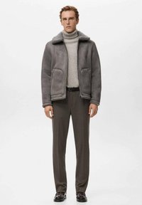 Grijze shearling jas met een kraag, gedragen over een lichtgrijze geribde coltrui, gecombineerd met donkergrijze, op maat gemaakte broek en zwarte schoenen.