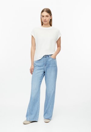 Vrouw staat en draagt een wit topje met korte mouwen, lichtblauwe wijde jeans en beige schoenen, met één hand in de zak tegen een witte achtergrond.