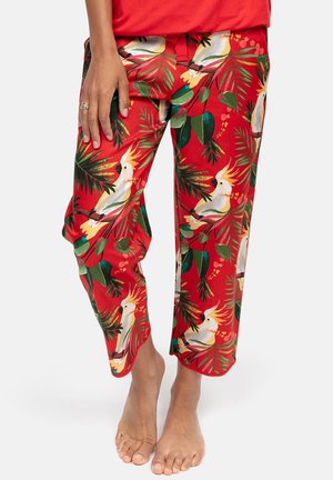 Czerwone spodnie z kolorowym tropikalnym nadrukiem liści i białego kakadu, noszone przez boso stojącą osobę.