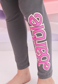 Grå leggings med en åtsittande design, med stora rosa och vita "Barbie"-bokstäver på vänster ben. Mjukt, elastiskt bomullstyg med rena fållar.
