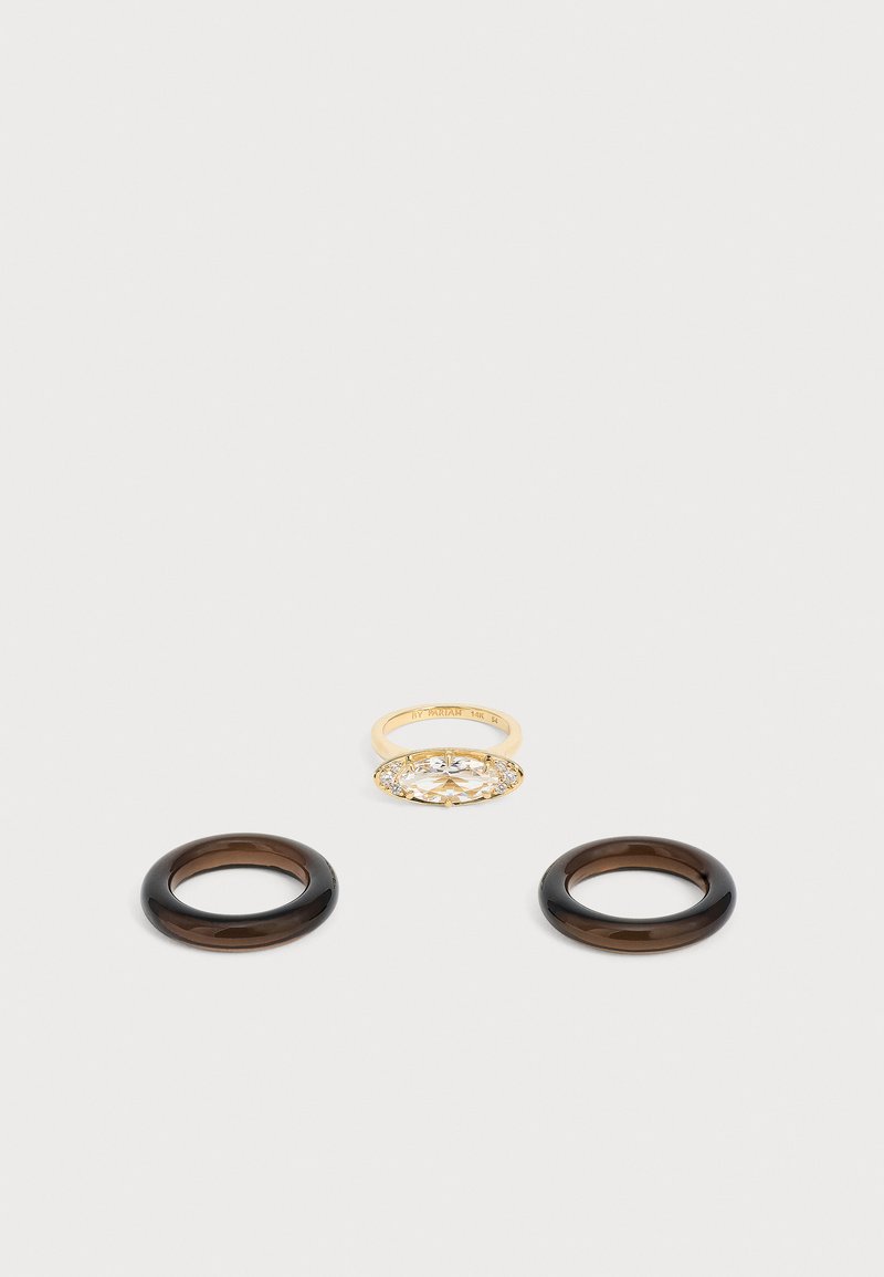 Trois bagues sur une surface claire : une bague en or avec une grande pierre ovale transparente entourée de petites pierres, et deux bagues circulaires unies de couleur brun foncé.