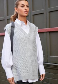 Gilet en tricot gris clair avec un col en V, présentant des motifs en torsades et texturés. Porté sur une chemise blanche à boutons, affichant un style superposé.