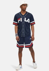 Maglia blu navy con logo Fila bianco, chiusura a bottoni e maniche a righe. Abbinata a pantaloni corti blu navy con dettagli rossi e bianchi. Cappello a secchiello bianco.