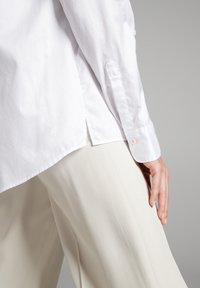 Chemise blanche à manches longues avec une texture nette, dotée d'un poignet à bouton unique et d'une fente sur le côté. Associée à un pantalon plissé beige clair.