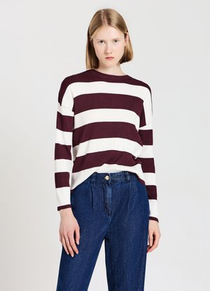 Donna con capelli lisci biondi che indossa un maglione a righe bordeaux e bianche a maniche lunghe e jeans blu a vita alta, in piedi davanti a uno sfondo semplice.