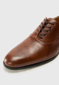 Zapato de cuero marrón, con un diseño de punta afilada, cordones oscuros en contraste y una textura lisa con una suela de goma negra.